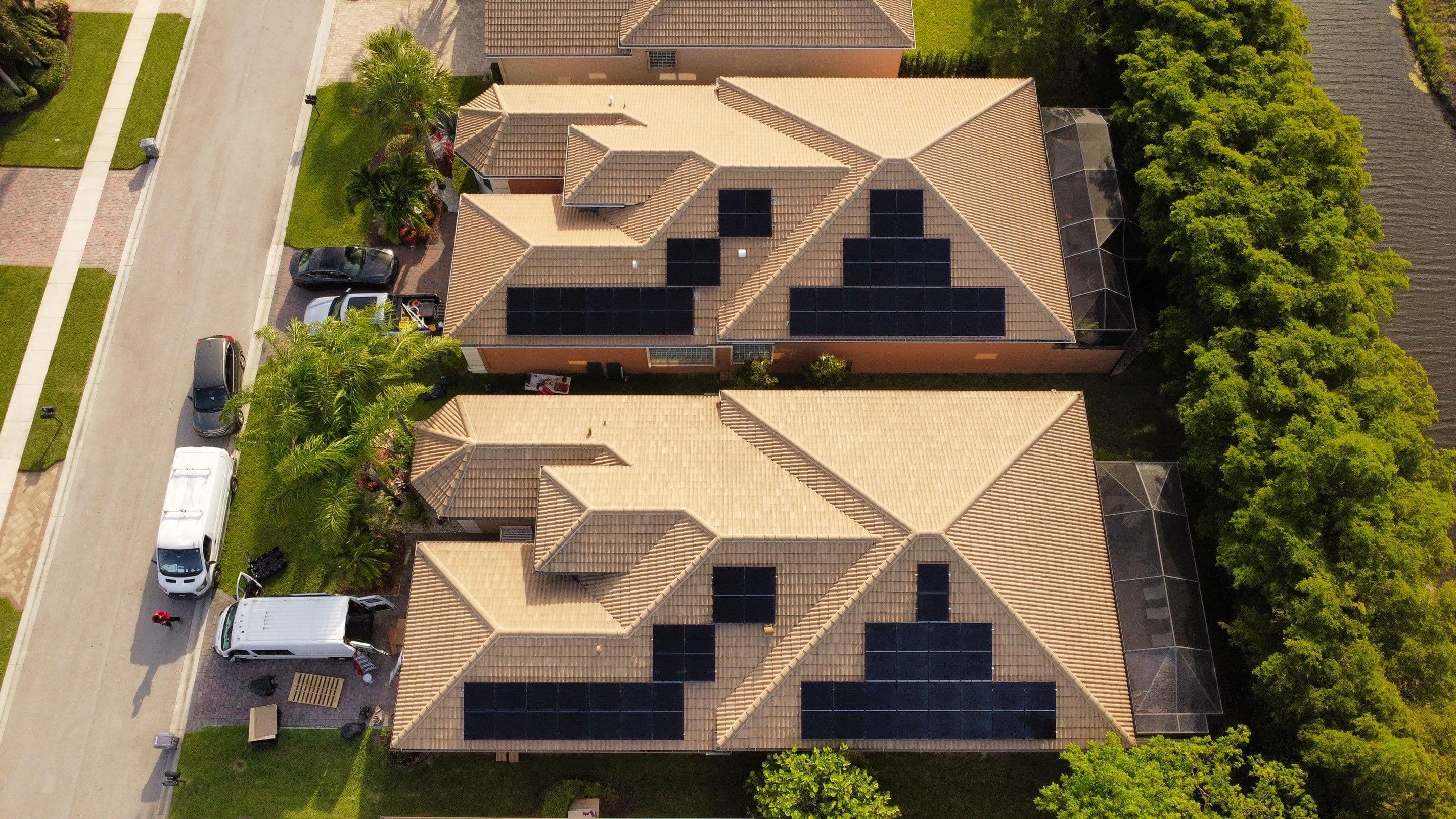 Aerial view of two suburban houses with solar panels on tan tile roofs and trees.