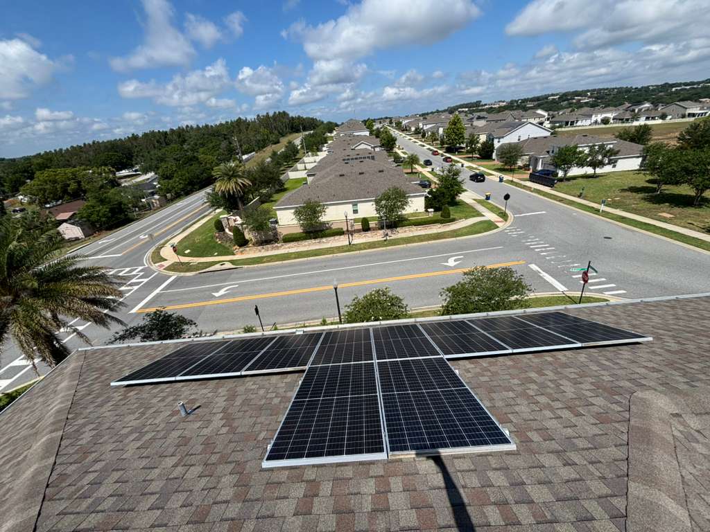 Solar panels on a shingled roof overlooking a suburban neighborhood under a bright blue sky.