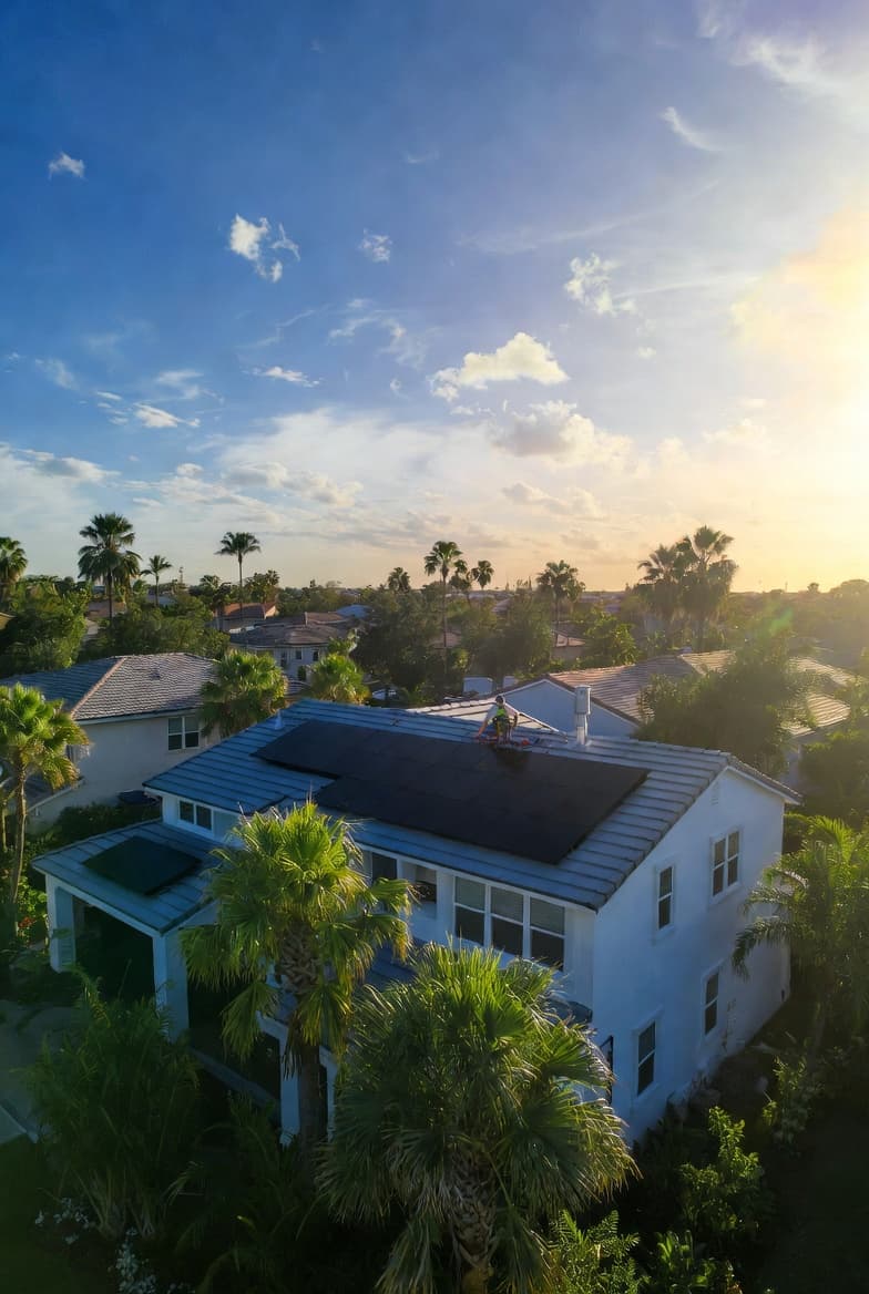 Solar panels on a white house roof surrounded by palm trees under a golden sunset.